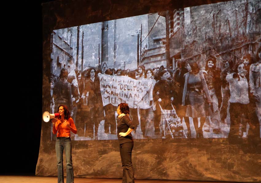 event image:Two women on stage and, behind them, a projection of a demonstration